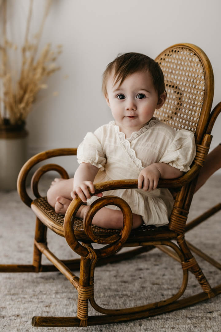 baby girls sits in a socking chair and looks towards the camera smiling during a baby photography session in st albans