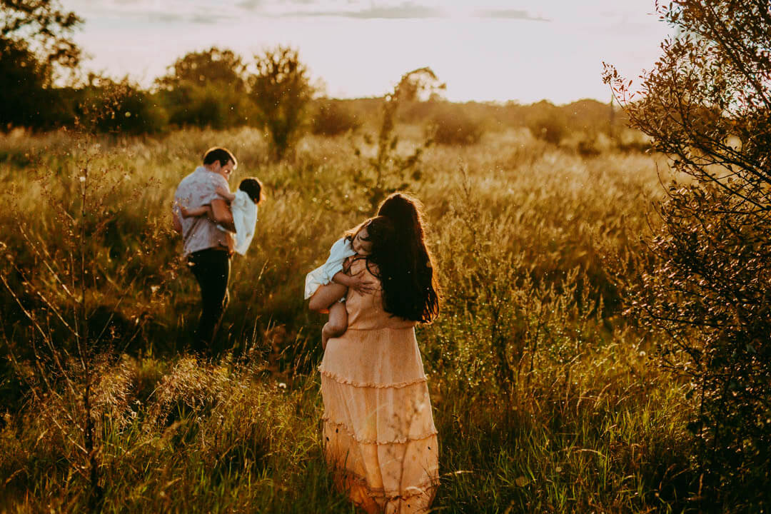 both parents carry a child and walk away while the sun sets in a field with long grass